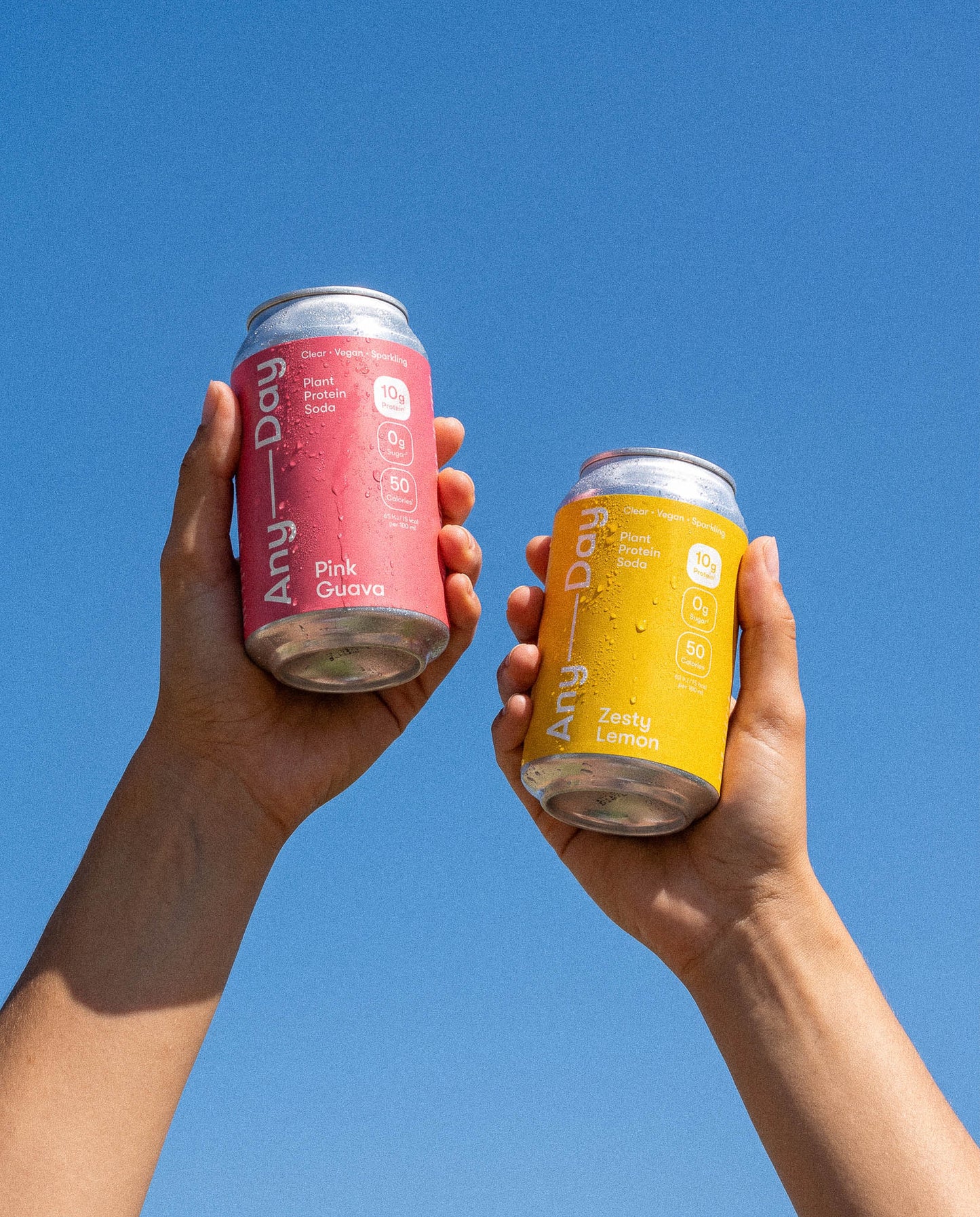 Zwei Hände halten pinke Anyday Pink Guava Dose und gelbe Anyday Zesty Lemon Dose vor blauem Himmel